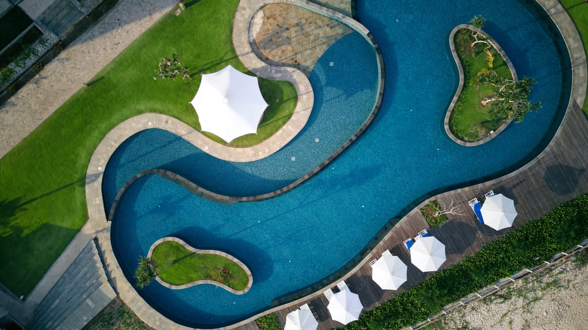Aerial view of a curved freeform lagoon pool with planted islands at a beachfront resort in Sumbawa